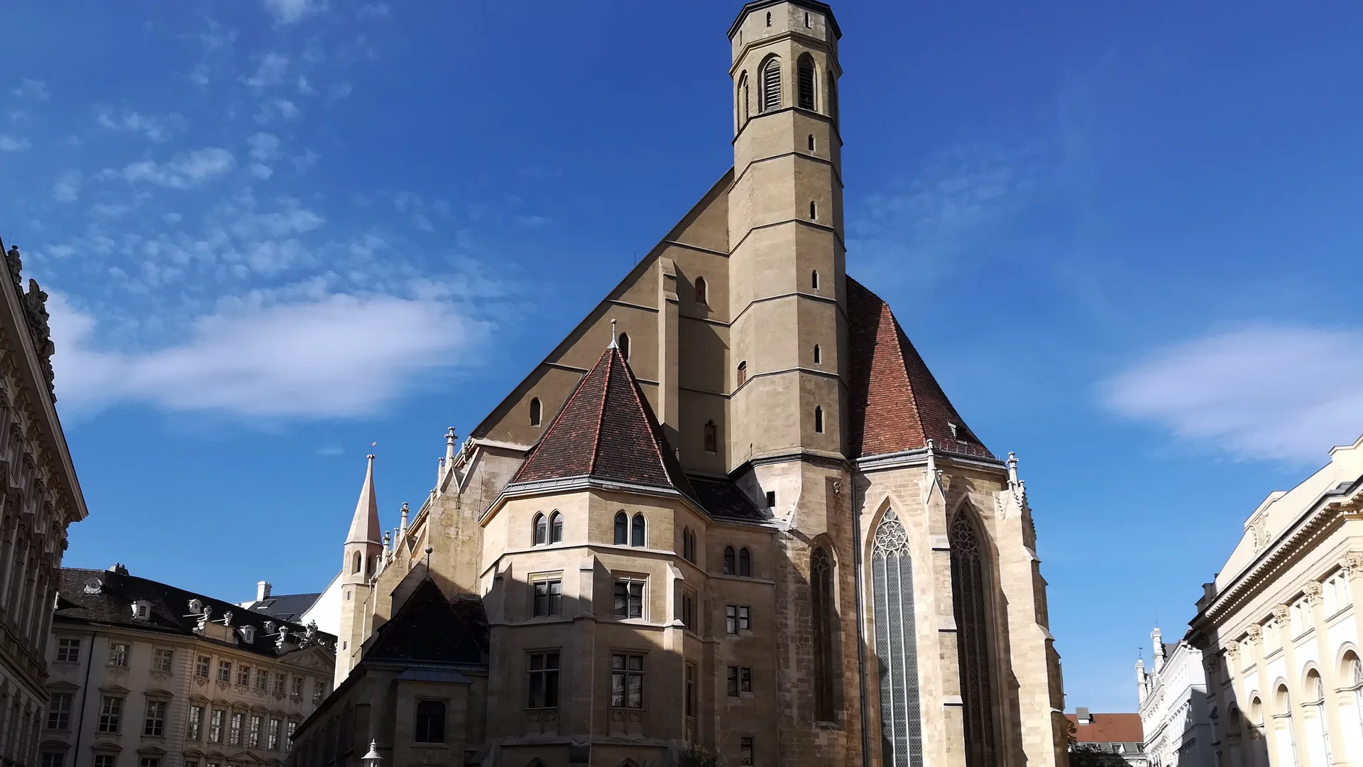 The Minoritenkirche in Vienna seen from Minoritenplatz: a pale-stone Gothic church with a tall slender tower, steep red-tiled roofs and tall traceried windows under a deep blue sky.