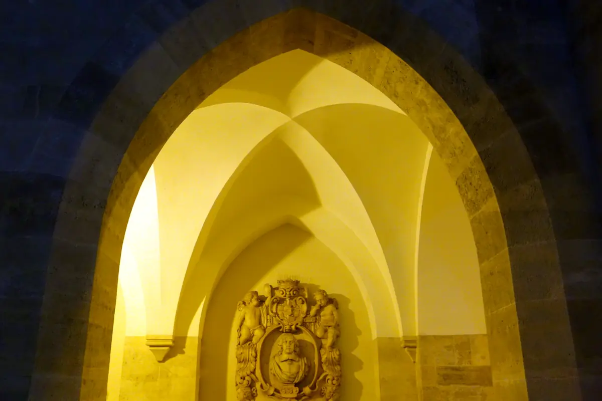 A pointed stone arch in the arcade of the Minoritenkirche glowing yellow at night, with ribbed vaulting and a carved coat of arms on the wall inside